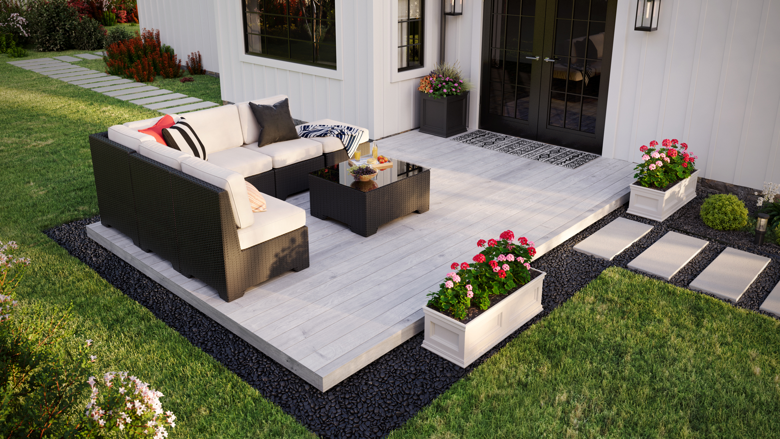 From a slightly raised angle, the image shows a deck attached to a white house. There is furniture in the left corner and a flower pot on the right. The deck sits atop gravel surrounded by grass.