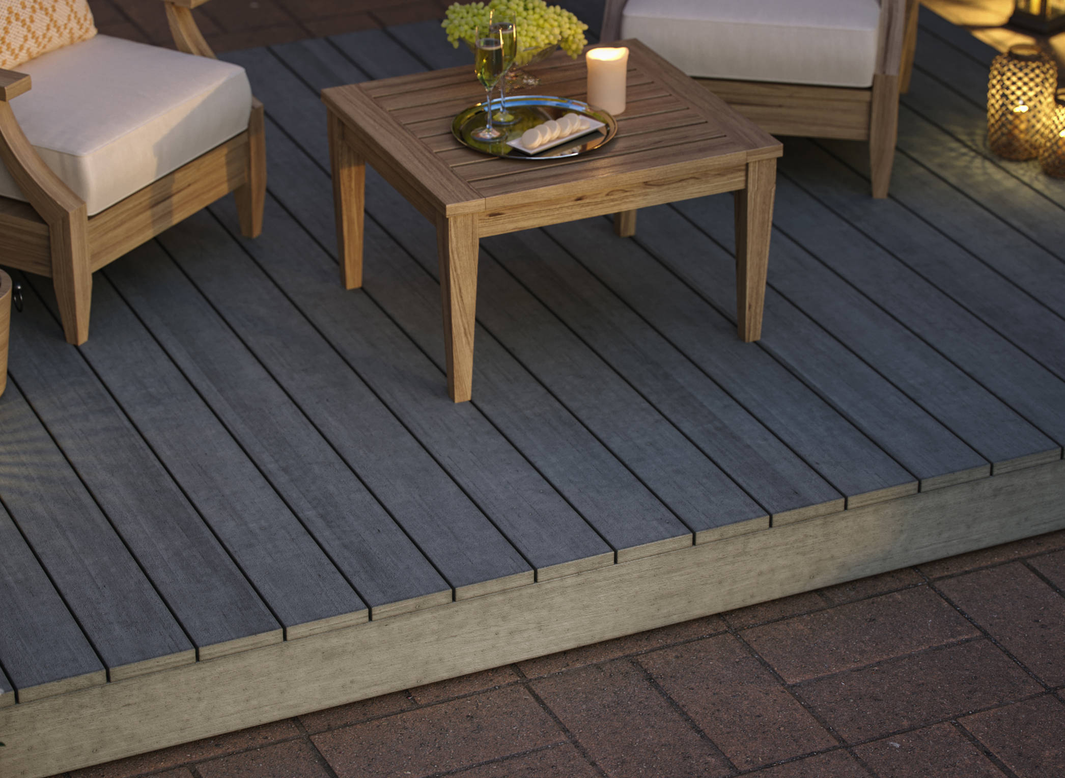 The image shows a deck with two chairs and a table. There is a tray, candle and centerpiece on the table. There are lanterns, champagne and flowers off to the sides, on the ground. The deck is on top of brick floors.