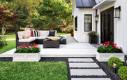 The image above shows an outdoor patio attached to a white house. The white deck holds a sofa and table. It sits atop gravel surrounded by grass.