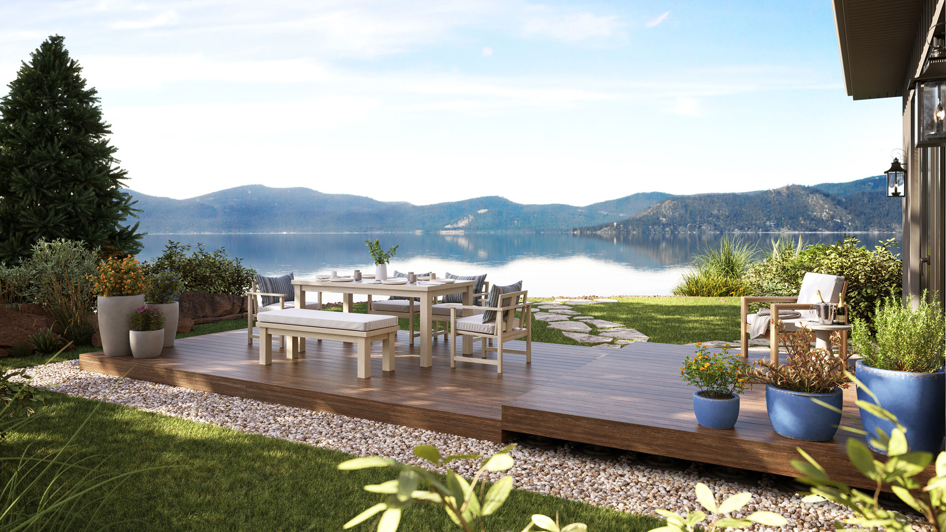There is a patio deck looking out towards a lake in the image above. There is a seating area along with plants, on the deck. There is grass and trees surrounding the area.