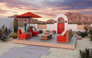 An outdoor patio is shown in this image with orange-cushioned furniture on top of it. There is a path to the deck with the seating area. The patio looks out towards mountains There are cacti and boulders beside the walls of the patio.